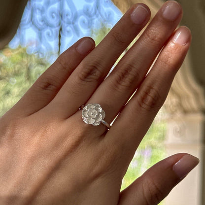 An Elagent Great Quality silver-toned ring with a flower-shaped clear gemstone is shown worn on a person's ring finger. The background is a softly blurred outdoor scene with light blue sky and greenery.